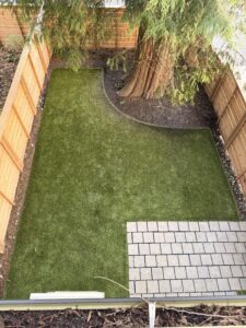 Small backyard with green artificial turf, curved garden bed around mature tree, wooden fencing, and paver patio