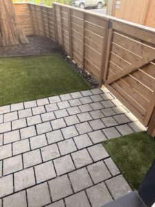 Backyard featuring artificial grass lawn, gray paver stone patio area, and natural wood horizontal fence